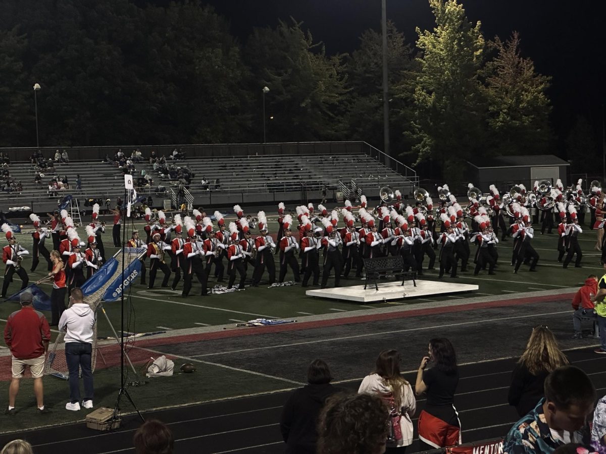 Fighting Cardinal Marching Band (FCMB) performing during halftime. Among the band members are Mentor’s 2025 Homecoming King and Queen, Benjamin Sherman (right) and Molly Tenney (left), both wearing gold sashes and crowns.